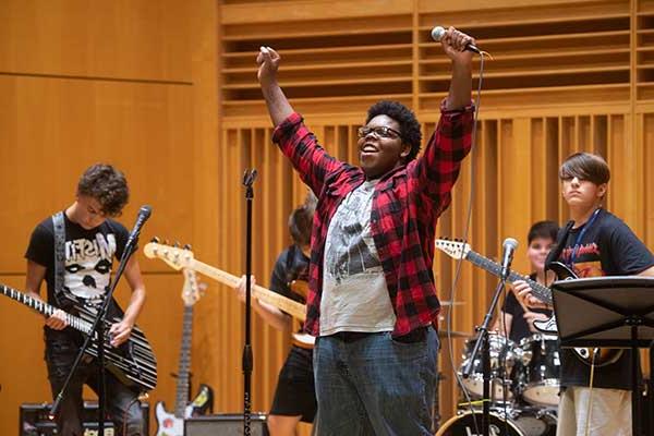 Five young men of mixed races perform music. In the front of the group is a Black man wearing a white T-shirt, a black-and-red plaid jacket, and blue jeans. He holds a microphone with his arms raised. Behind him are four males wearing black T-shirts. Three hold guitars and one sits behind a drum set.